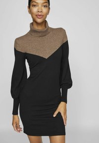 Abito a dolcevita nero e marrone con maniche lunghe a sbuffo, silhouette aderente e un motivo a chevron a contrasto sul petto. Tessuto a maglia morbida.