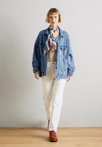 Giacca di denim con tasche frontali, indossata sopra una maglietta beige, abbinata a pantaloni bianchi e scarpe marroni. Una sciarpa multicolore aggiunge un tocco di dettaglio.