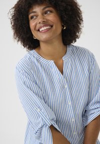 Chemise rayée bleu clair et blanche, avec un col rond, des boutons de fermeture et des manches retroussées agrémentées de boutons.