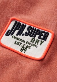 Korala koszulka z tkaniny z białą i pomarańczową metką oraz niebieskim i zielonym napisem: "JPH. SUPER DRY GENERAL GOODS, LOT. 54®".