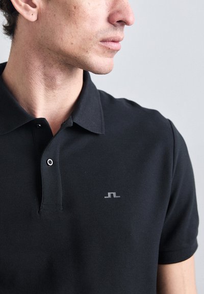 Polo negro hecho de una tela texturizada con una tapeta de tres botones y un pequeño logo gris en el lado izquierdo del pecho.