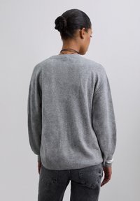 Žena s tmavými vlasy ve drdolu, oblečená v šedém svetru s dlouhým rukávem a tmavě šedých džínách, stojící zády k jednoduchému pozadí.