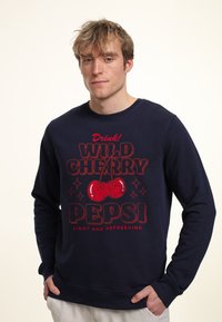Felpa navy con stampa grafica rossa che presenta la scritta "Wild Cherry Pepsi" e illustrazioni di ciliegie. Maniche a costine e scollo rotondo. Tessuto morbido.