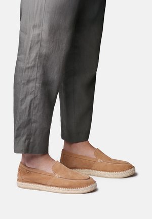 Person, die graue, gekürzte Hose und hellbraune Wildleder-Espadrille-Slipper mit geflochtenen Jutesohlen trägt und auf einem schlichten hellen Hintergrund steht.