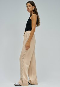 Beige wijde broek met een tailleband met koord en een tonale ruitpatroon langs de zijkant. Gecombineerd met een zwart mouwloos topje en sportieve schoenen.