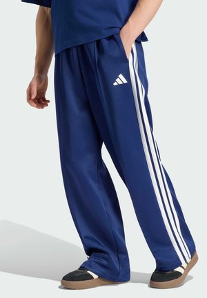 Osoba ubrana w niebieskie spodnie dresowe Adidas z białymi paskami i czarne buty z gumowymi podeszwami, stoi z jedną ręką w kieszeni.