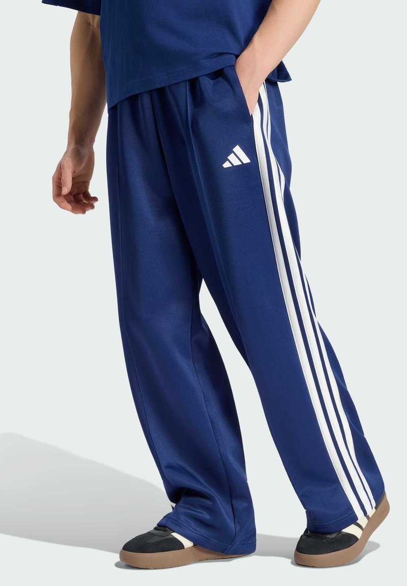 Personne portant un pantalon de survêtement Adidas bleu avec des bandes blanches et des chaussures noires à semelles en caoutchouc, debout avec une main dans la poche.