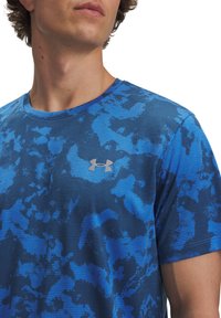 Camiseta atlética azul con un patrón texturizado y ondulado. Logo plateado de Under Armour en el pecho. Cuello redondo y mangas cortas.