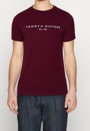 Homme portant un t-shirt bordeaux Tommy Hilfiger avec logo et inscription "EST. 1985", associé à un pantalon bleu foncé, debout devant un fond uni.