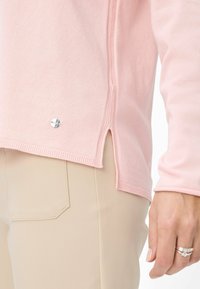 Lichtroze trui met ribbelstructuur, zijsplitten aan de onderkant en een klein zilveren logo accent. Gecombineerd met beige broek.