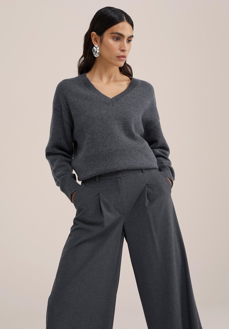 Grijze wolblend trui met een V-hals, geribbelde manchetten, gecombineerd met grijze wijde broek met plooien aan de voorkant. Model kijkt recht vooruit.