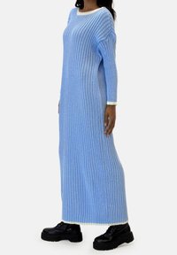 Robe en maille côtelée bleu clair avec manches longues et col blanc, présentant une coupe ample et une longueur jusqu'aux chevilles. Portée avec des bottes noires.