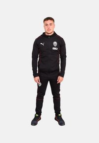 Chándal negro hecho de tela suave, con detalles en rojo y un logo en el pecho y los pantalones. Combinado con zapatillas deportivas negras.