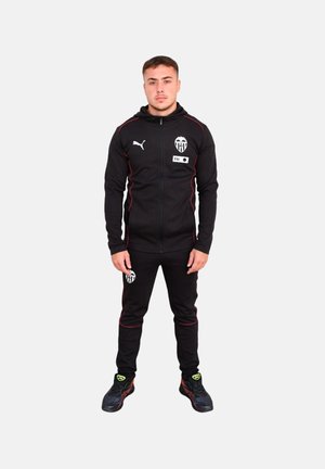 Chándal negro hecho de tela suave, con detalles en rojo y un logo en el pecho y los pantalones. Combinado con zapatillas deportivas negras.