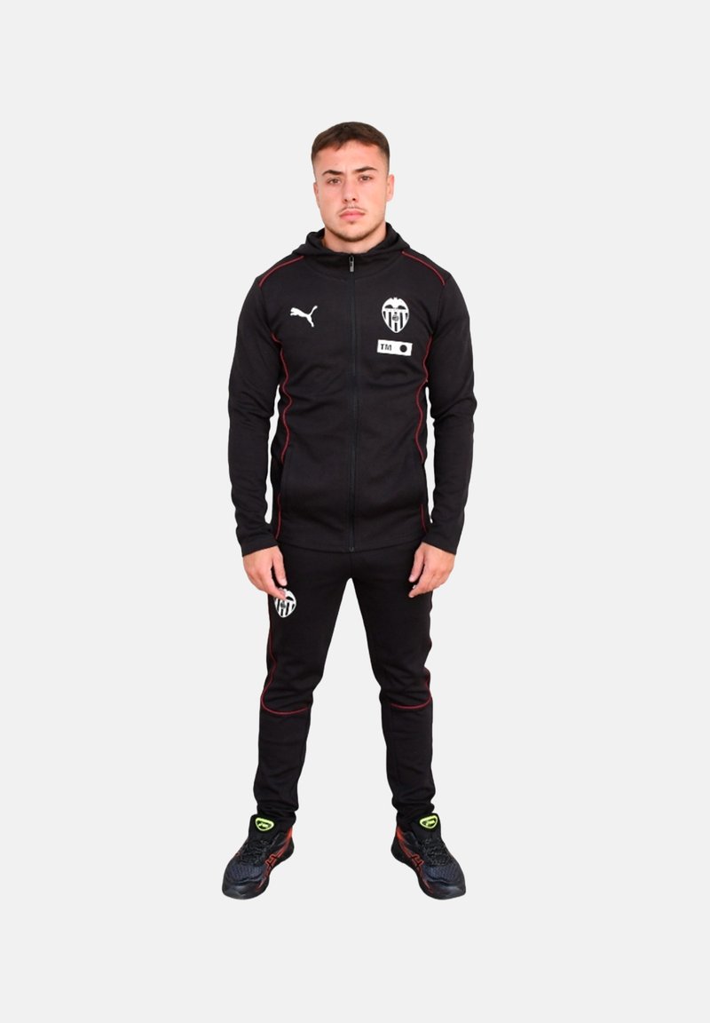 Chándal negro hecho de tela suave, con detalles en rojo y un logo en el pecho y los pantalones. Combinado con zapatillas deportivas negras.