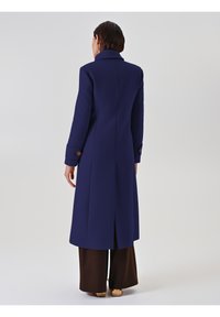 Lungo cappotto blu navy con colletto, spacco posteriore e polsini con bottoni, realizzato in un tessuto liscio. Abbinato a pantaloni larghi marroni.