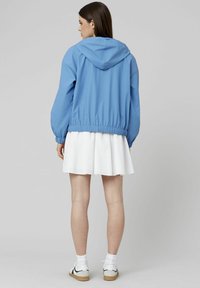 Veste à capuche bleu clair avec des poignets et un ourlet élastiques. Portée par-dessus une jupe blanche et associée à des chaussettes blanches et des chaussures claires.