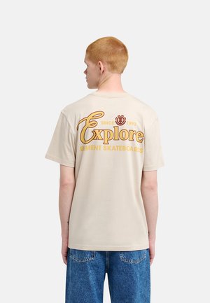Jong persoon met kort rood haar die een beige T-shirt met de tekst "Explore Element Skateboards" en een blauwe spijkerbroek draagt, met de rug naar de camera toe.