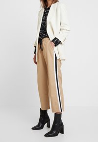 Blazer crème, chemise noire à motifs, pantalon de jogging tan avec des rayures noires et blanches, et bottines noires à talon large.