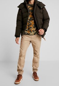 Sort puffjakke med hætte, camouflage-mønstret t-shirt i grønne og brune nuancer, beige bukser og brune høje sneakers med hvide såler.