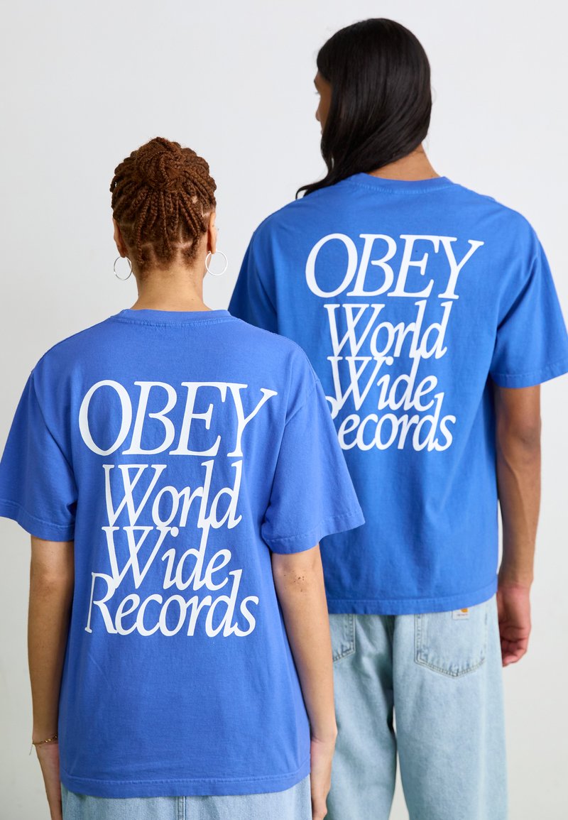 Blå bomull t-skjorter med avslappet passform har stor hvit tekst på baksiden som sier "OBEY World Wide Records." Enkle crew neck-design.