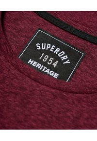Camiseta burdeos con una etiqueta negra que lleva un texto blanco que dice "SUPERDRY 1954 HERITAGE". Tejido de algodón con un aspecto texturizado.