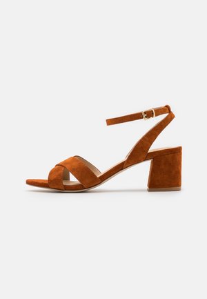 Brune ruskinds hælede sandaler med åben tå og tværgående remdesign. Har en ankelrem med en guldbuckle og en blokhælt.