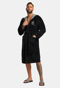 Schwarzer, plüschiger Bademantel mit grauem Kapuzen, seitlichen Taschen und einem Bindegürtel. Mit einem gestickten Logo auf der Brust und einer weichen Textur.