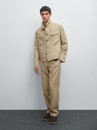 Beige, strukturierte Jacke mit hohem Kragen, zwei Brusttaschen und braunen Knöpfen; kombiniert mit passenden weit geschnittenen Hosen und dunklen Schuhen.