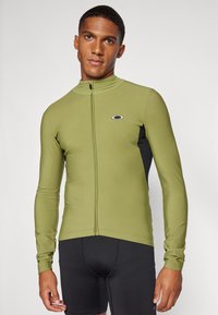 Haut de sport à manches longues en vert olive avec un panneau noir, doté d'un col haut et d'une fermeture éclair sur le devant, fabriqué dans un tissu lisse et léger.
