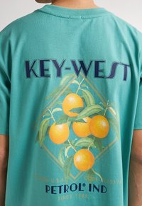 Turkoosi puuvillapaita, jossa on grafiikka appelsiineista ja teksti "KEY-WEST" sinisellä, vihreillä lehdillä ja kultaisilla korostuksilla.