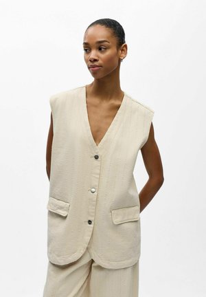 Woman wearing a sleeveless beige buttoned vest with two front pockets and matching pants, standing against a plain background.