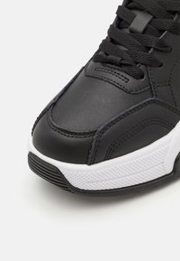 Zapato deportivo negro con parte superior de cuero suave, acentos de plástico, cordones negros, entresuela blanca texturizada y suela de goma con agarre.