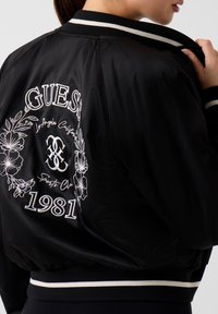 Giacca bomber in raso nero con ricamo floreale bianco, con scritte "GUES" e "1981", collo a costine e dettagli a righe.