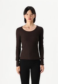 Brauner langärmeliger Ripp-Top mit Rundhalsausschnitt, enganliegendem Design und strukturiertem Stoff, kombiniert mit schwarzen Hosen, vor einem weißen Hintergrund präsentiert.