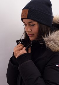 Black parka jacket with fur-lined hood, zippered front, ribbed cuffs, and a leather patch on a navy beanie. Model is adjusting the jacket.