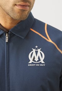 Mornarska jakna iz blaga z zadrgo, ki ima oranžne šive in bel logo s tekstom "DROIT AU BUT" na predelu prsi.