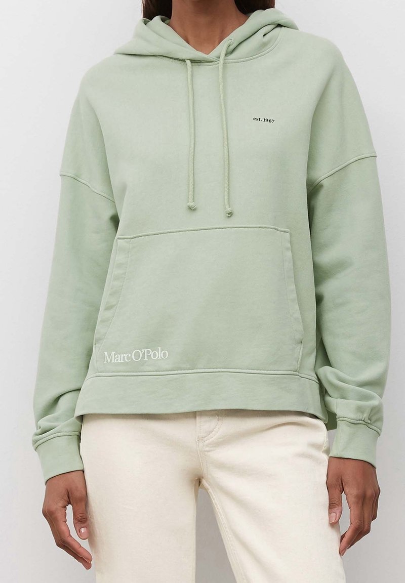 Personne portant un sweat à capuche vert clair avec poche kangourou à l'avant et pantalon crème, texte « est. 1967 » sur la poitrine et « Marc O'Polo » près de la poche.