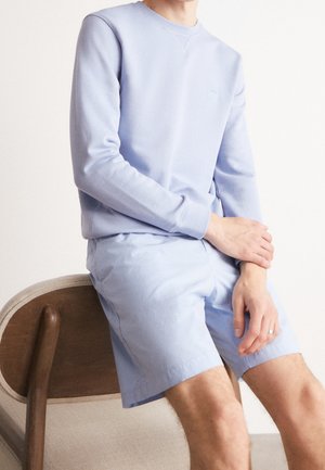 Person sitzt auf einem Holzstuhl, trägt ein hellblaues Langarmhemd und passende Shorts, die Hände ruhen auf einem Knie.