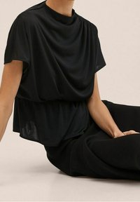 Top negro holgado con un cuello alto, tejido drapeado y mangas cortas. Combinado con pantalones negros que destacan por su textura suave.