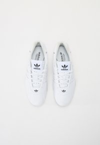 Fehér sportcipők sima bőrből, fűzős kialakítással és fekete Adidas logóval a nyelvén és az oldalán.