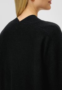 Schwarzer Strickpullover mit einem strukturierten Muster, V-Ausschnitt hinten und lockerer Passform. Der Stoff wirkt weich und leicht dick.