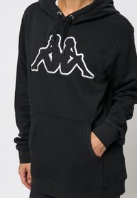 Zwarte hoodie van katoen met een voorste kangaroozak. Het ontwerp bevat een witte geborduurde graphic van twee zittende figuren.