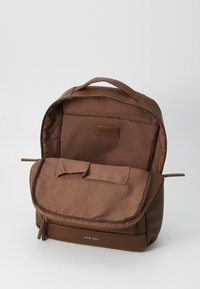 Mochila marrón con un exterior suave, cierre de cremallera y una solapa frontal. En el interior tiene un forro de tela y un bolsillo adicional para organización.