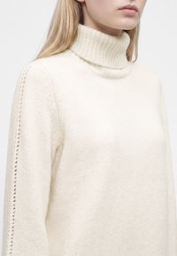 Femme portant un pull à col roulé côtelé de couleur crème avec un détail décoratif en maille ajourée le long des manches.