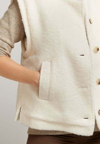 Gilet en polaire de couleur blanc crémeux avec une texture douce, doté d'une fermeture à boutons, de deux poches latérales et d'une coupe décontractée avec fentes sur les côtés.