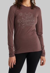 Hgrunnjakki með langum ermum í brúnu, úr mjúku efni, með hvítu grafíkinu "Færðu þig náttúrulega" í miðjunni á framan.