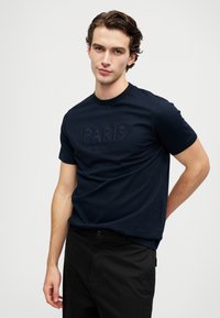 Tumši zilā kokvilnas T-krekls ar teksturētu izšūtu "PARIS" logo un papildus tekstu. Īsas piedurknes, apaļa kakla izgriezums, gluda auduma.