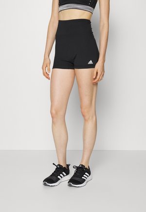 Woman wearing black high-waisted Adidas shorts and black Adidas sneakers standing against a plain light background.