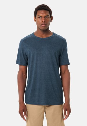 Uomo in piedi, vista frontale, che indossa una maglietta a maniche corte blu scuro semplice e pantaloni beige chiaro su uno sfondo bianco.